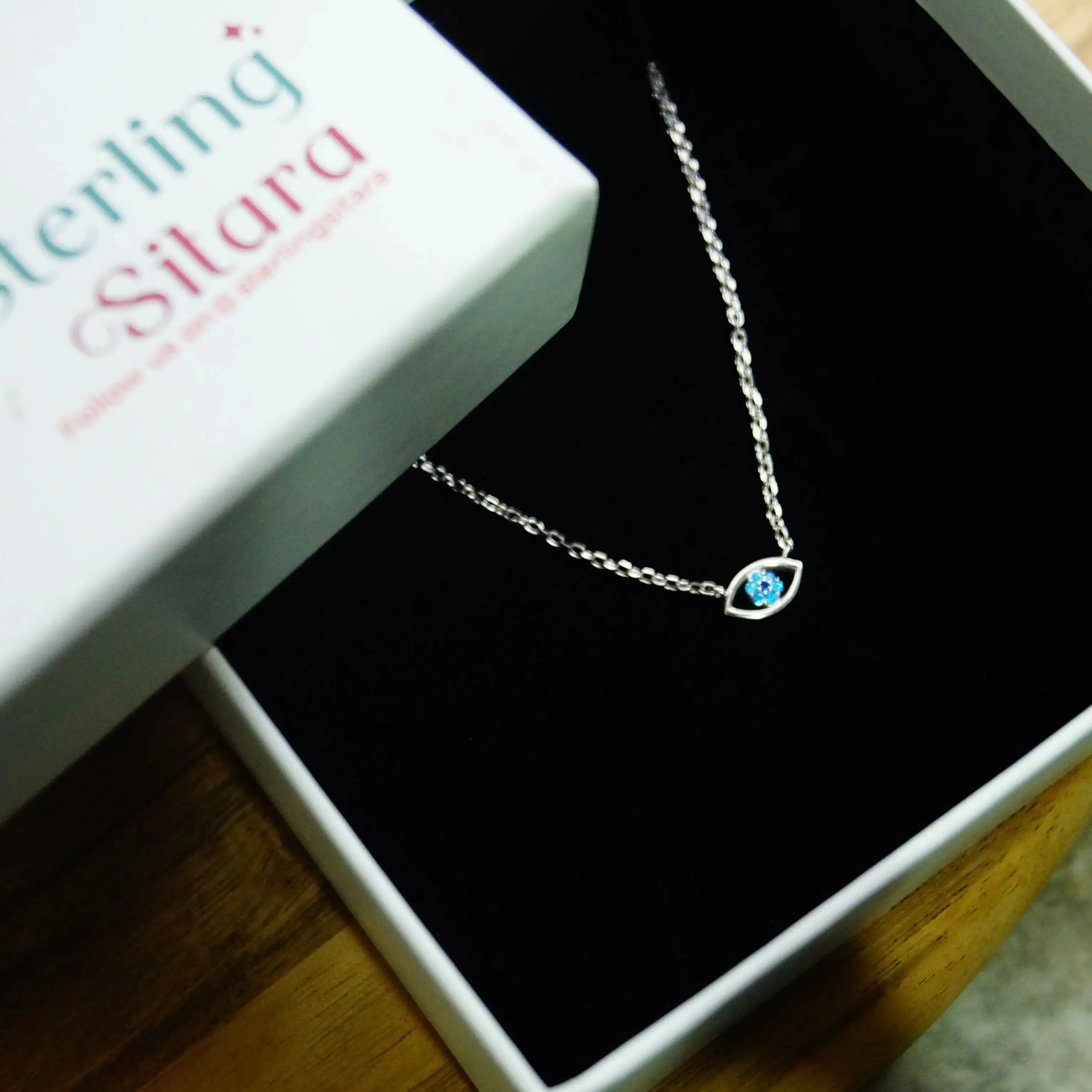 Silver necklace with a blue stone on a black box, branded 'Sterling Silana', with a plant in the background.